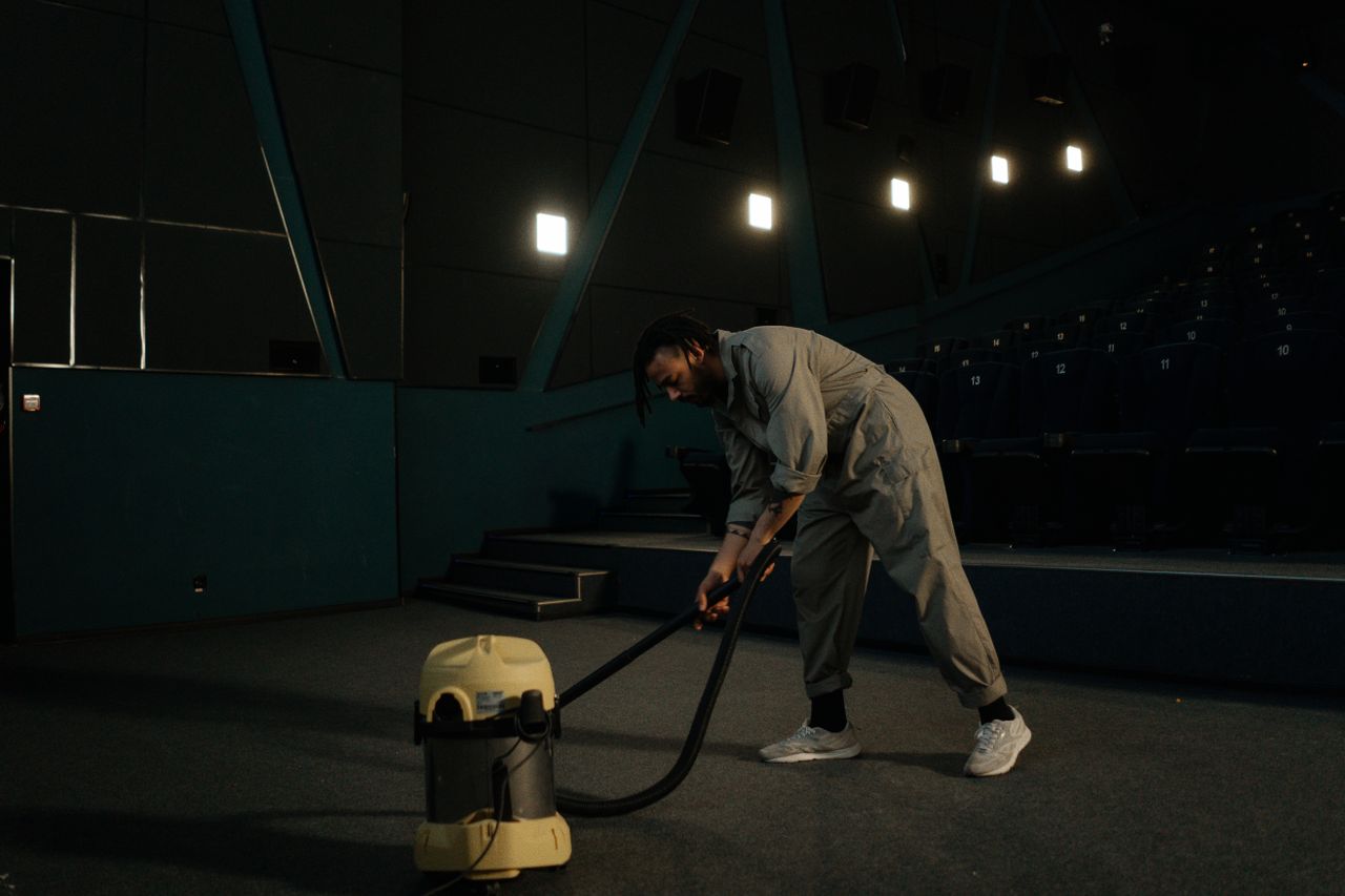 A movie theater worker is vacuuming the auditorium floor beneath low lighting.