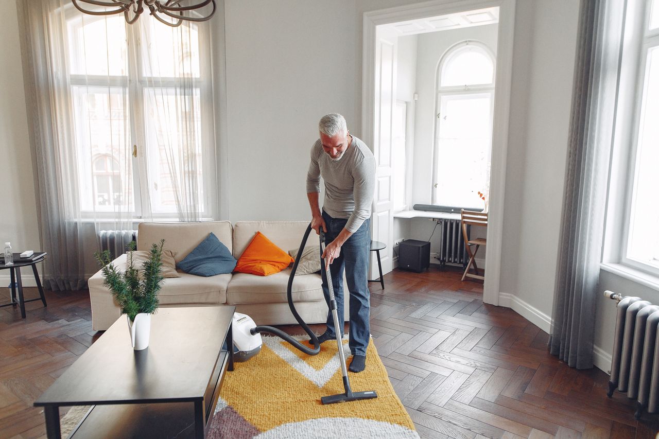 Seen from above, a focused grey-haired man with a beard is vacuuming the carpet at home